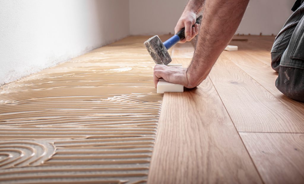 Ein Bodenleger verlegt Holzparkett in einem Raum, während er mit einem Gummihammer und einem Abstandshalter arbeitet.